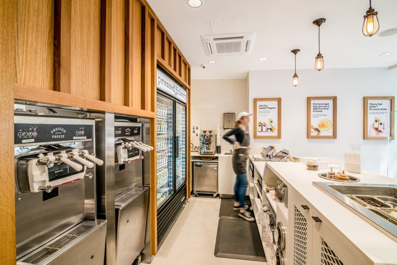 Pressed Juicery service counter with built-in soft-serve machines, a glass-front beverage cooler, wood paneling, and pendant lighting Source: Blueberry Builders