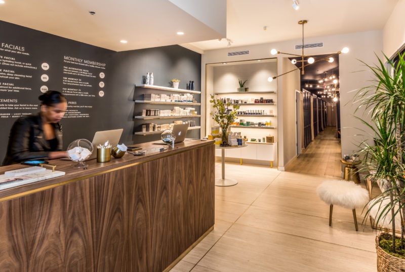 Reception area of a modern spa with a wooden desk, a staff member at a laptop, product shelves, plants, and a hallway leading to treatment rooms.