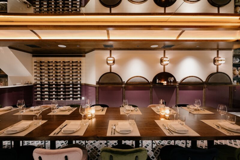 L’Artusi dining room with a long wood dining table, colorful chairs, patterned tile floors, arched wall details, and warm illuminated ceiling accents Source: Blueberry Builders